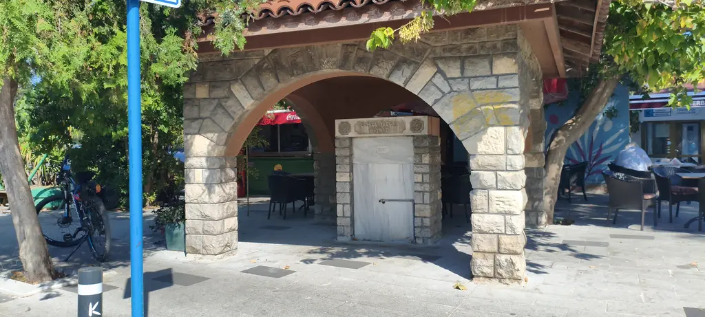 Fenerbahçe Parkı'nda çeşme; kesme taştan kemerli çatı, ortada beyaz mermer ayna taşı ve arkada kafe masaları.