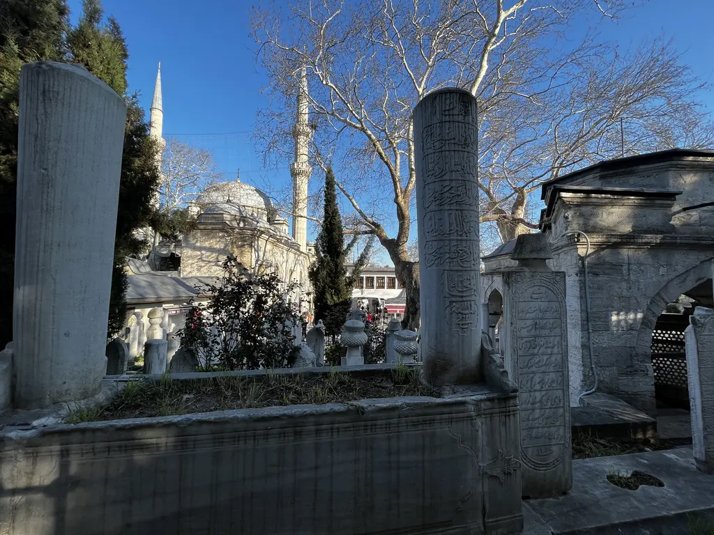 Eyüp Sultan Camii çevresinde hazire; yüksek mermer mezar taşları, ağaçlar ve arkada caminin kubbesiyle minaresi.