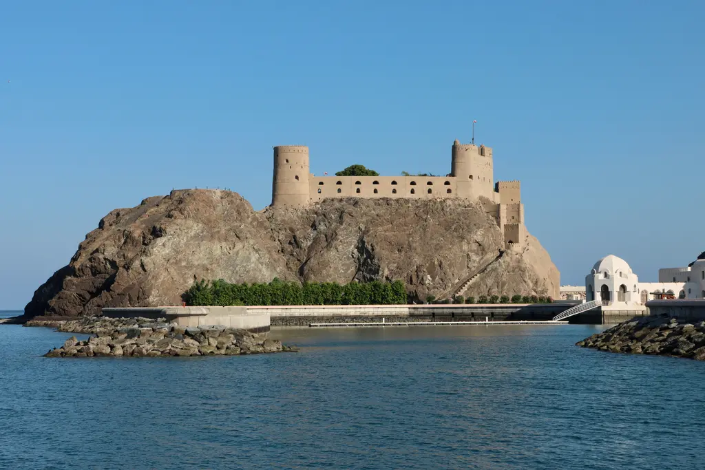 Maskat'ta kayalık bir burnun tepesine kurulmuş bej taş kale; önünde turkuaz deniz, solda çıplak kayalar ve sağda beyaz kubbeli yapı ile liman rıhtımı.