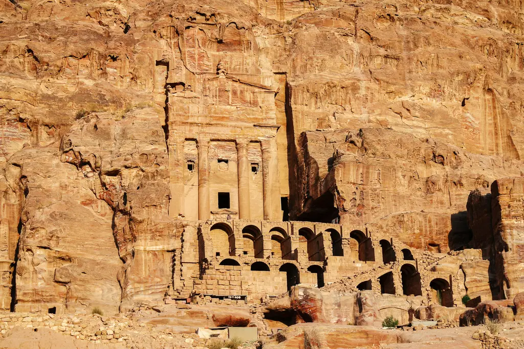 Petra'da kayaya oyulmuş anıtsal mezar cephesi; sütunlu üst kat, kapı boşlukları ve altta sıralanan kemerli taş odalar, çevresinde çizgili kumtaşı kayalıklar.