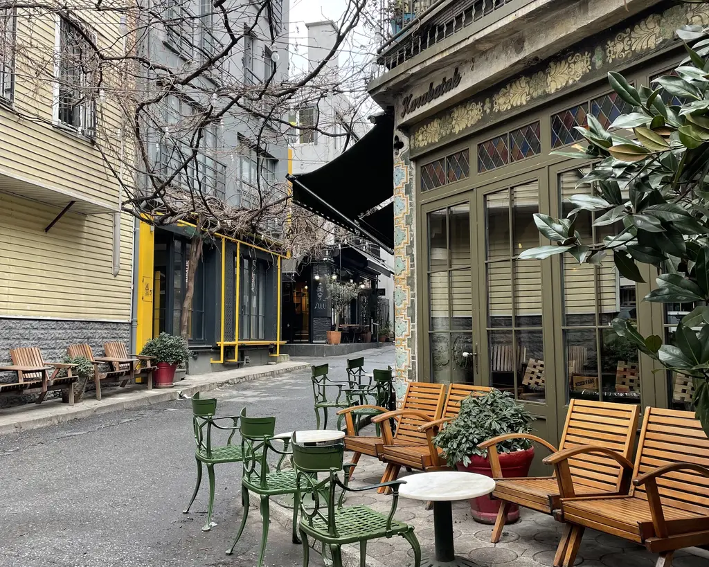 Karaköy'de bir kafe sokağı; ahşap sandalyeler ve yeşil metal masalar, saksılarda bitkiler ve yaprağını dökmüş ağaç.