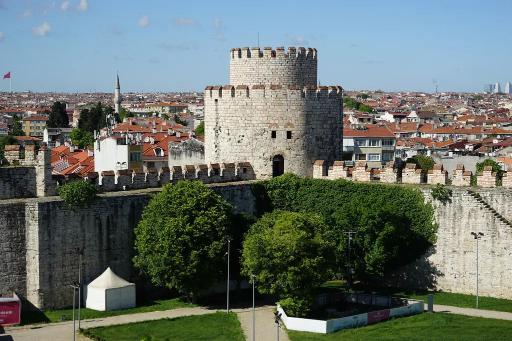 Yedikule Hisarı'nda kırmızı kiremitli mazgallı büyük yuvarlak kule yüksekten; önde mazgallı duvar ile ağaçlar, arkada kırmızı çatılı mahalle ve bir cami minaresi.