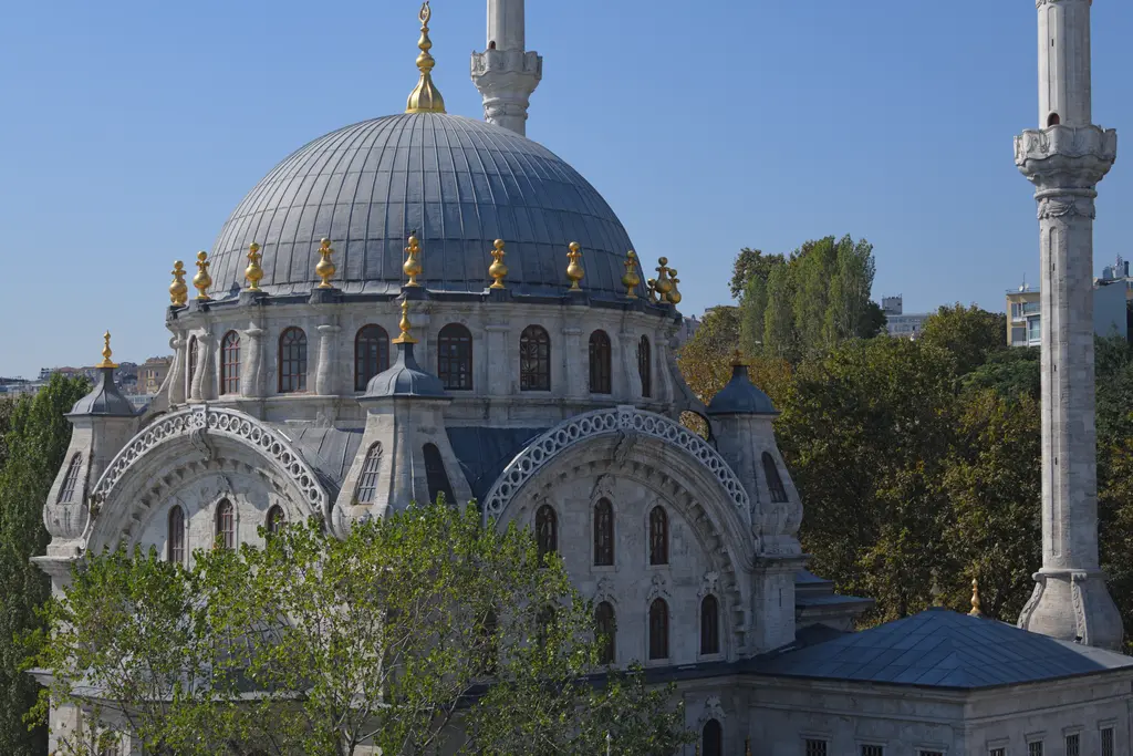 Nusretiye Camii; mavi gökyüzü altında kurşun kubbe, yaldızlı alemler, kemerli pencere sıraları, sağda ince yivli minare ve önde ağaçlar.