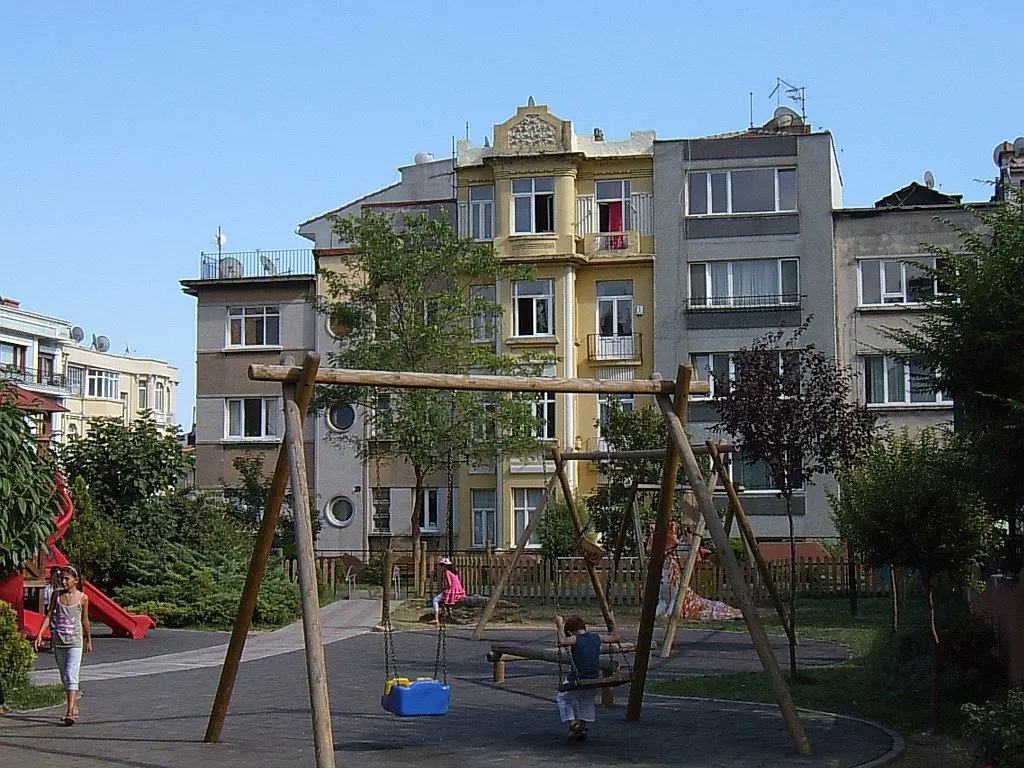 Cihangir Parkı'nda çocuk oyun alanı; ahşap salıncaklar, kaydırak, ağaçlar ve arkada balkonlu eski apartmanlar.