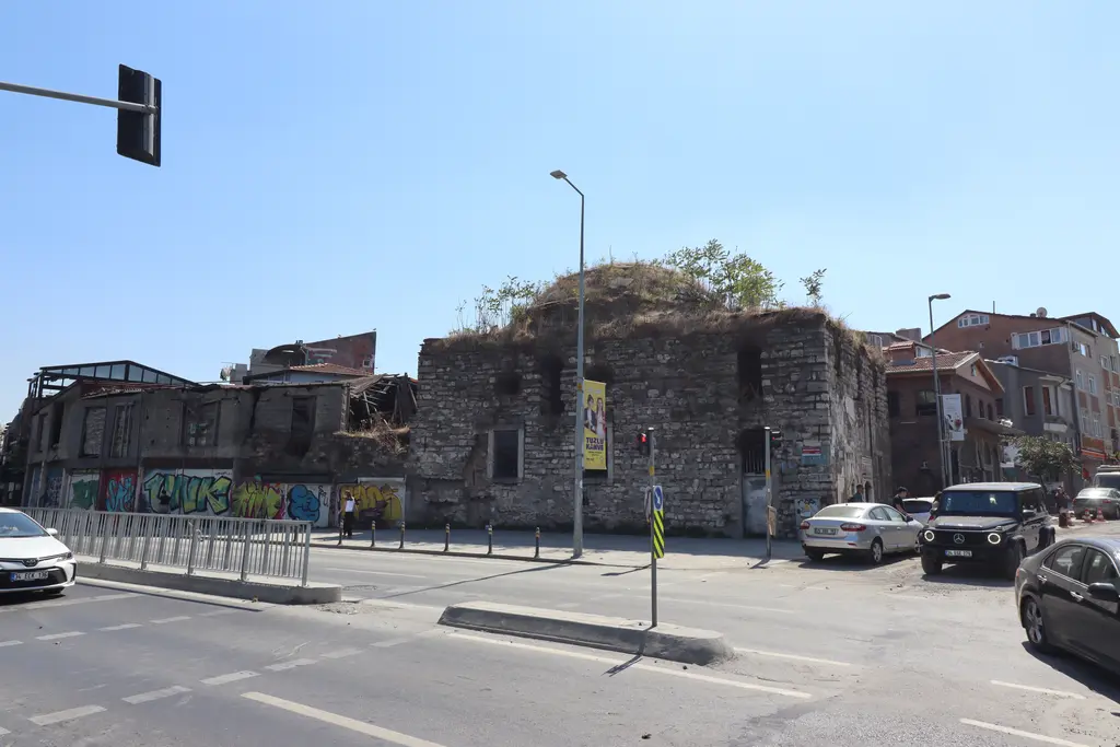 Ayakapı Hamamı'nın caddeden görünümü; harap taş gövde, çatısında otlar, önde trafik ışığı ve park etmiş arabalar.