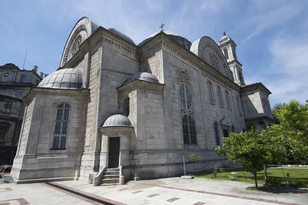 Aya Triada Kilisesi'nin yan cephesi; taş duvar, kemerli pencereler, yarım kubbeler, arkada çan kulesi ve önde bahçe.