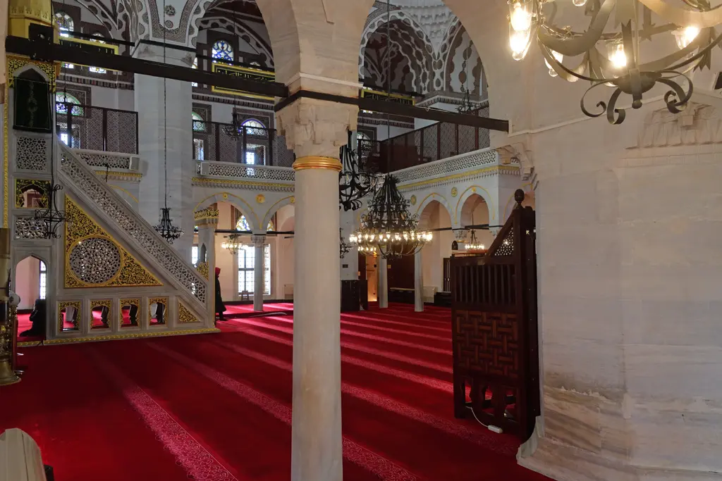 Yeni Valide Camii'nde yan mahfil; mermer sütunlar, yaldızlı minber, avizeler ve kırmızı halılı harim.