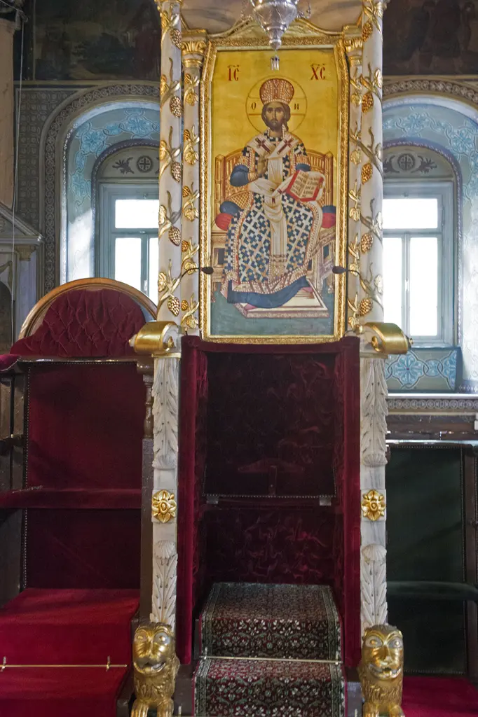 Yaldızlı ahşap piskopos tahtı; kırmızı kadife oturak, üstünde kubbeli baldaken, arkada aziz tasvirli ikona ve mermer duvar.