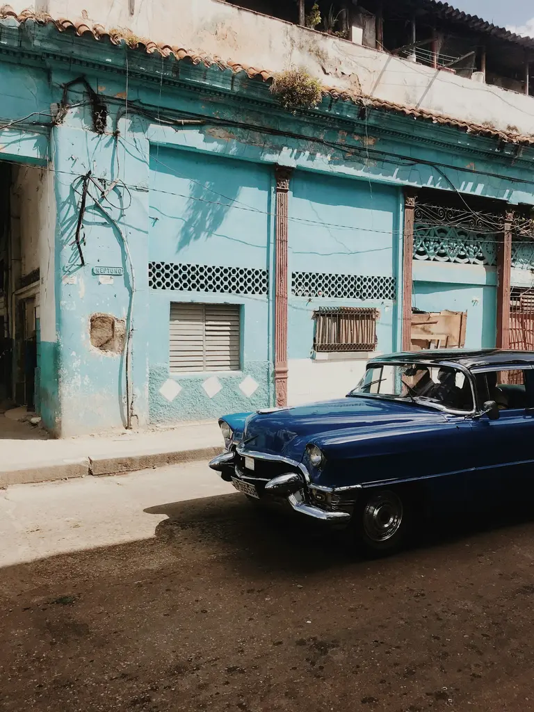 Küba'nın başkenti Havana'da turkuaz renkli, yıpranmış sömürge cephesinin önünde duran lacivert klasik Amerikan arabası; öğle güneşinde "Neptuno" sokak tabelası ve kiremitli saçak.