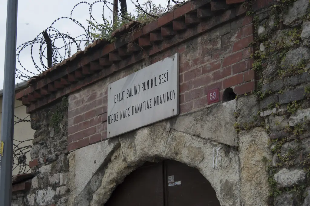 Panayia Balino Kilisesi'nin kapı levhası; tuğla duvar üzerinde Türkçe ve Rumca yazılı levha, altında taş kemer ve üstte dikenli tel.