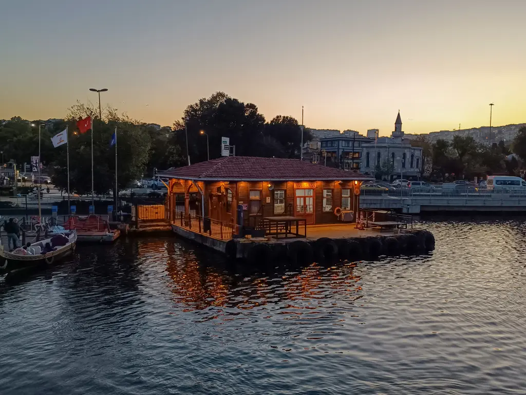 Eyüpsultan iskelesi; gün batımında ışıkları yanan ahşap iskele binası, Haliç'in suyu ve kıyıdaki tekneler.