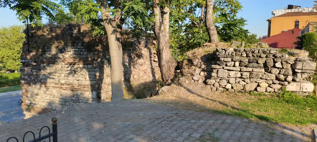 Haliç surlarından bir bölüm; ağaçların arasında yıkık taş duvar kalıntıları, önde taş döşeli yol ve uzakta sarı ev.