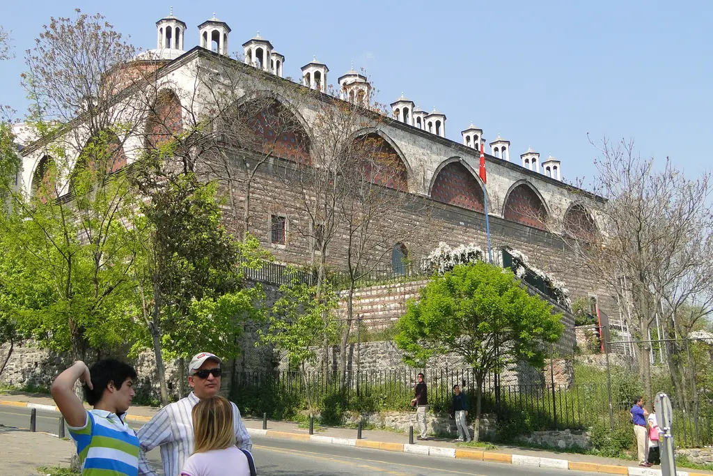 İlkbaharda Tophane-i Âmire; yeşillenen ağaçlar, kemerli taş cephe, çatıda kubbe fenerleri ve önde yürüyen insanlar.