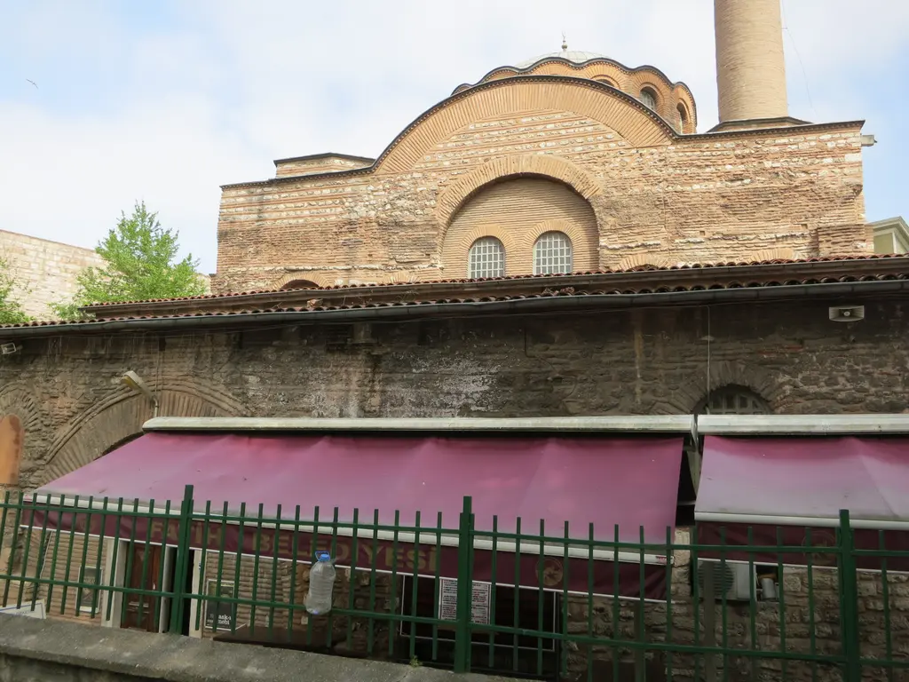 Kalenderhane Camii'nin yan cephesi; iki pencereli tuğla alınlık, arkada kubbe ve minare, önde bordo tenteler ile yeşil demir parmaklık.