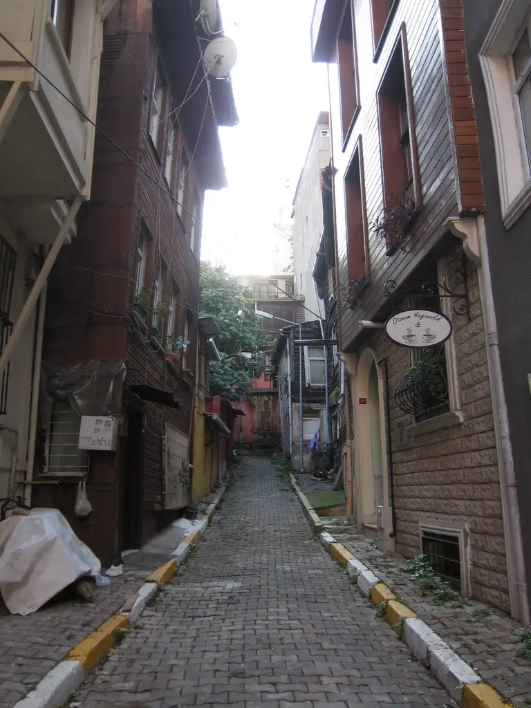 Eski ahşap evler arasında daralan arnavut kaldırımlı sokak; kenarlarda sarı çizgiler ve yeşillik.