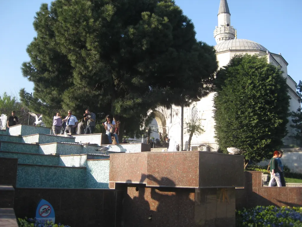 Sultanahmet Parkı'ndan Firuz Ağa Camii; önde basamaklı modern havuz ve granit bloklar, arkada ağaçlar arasından kubbe ile minare.