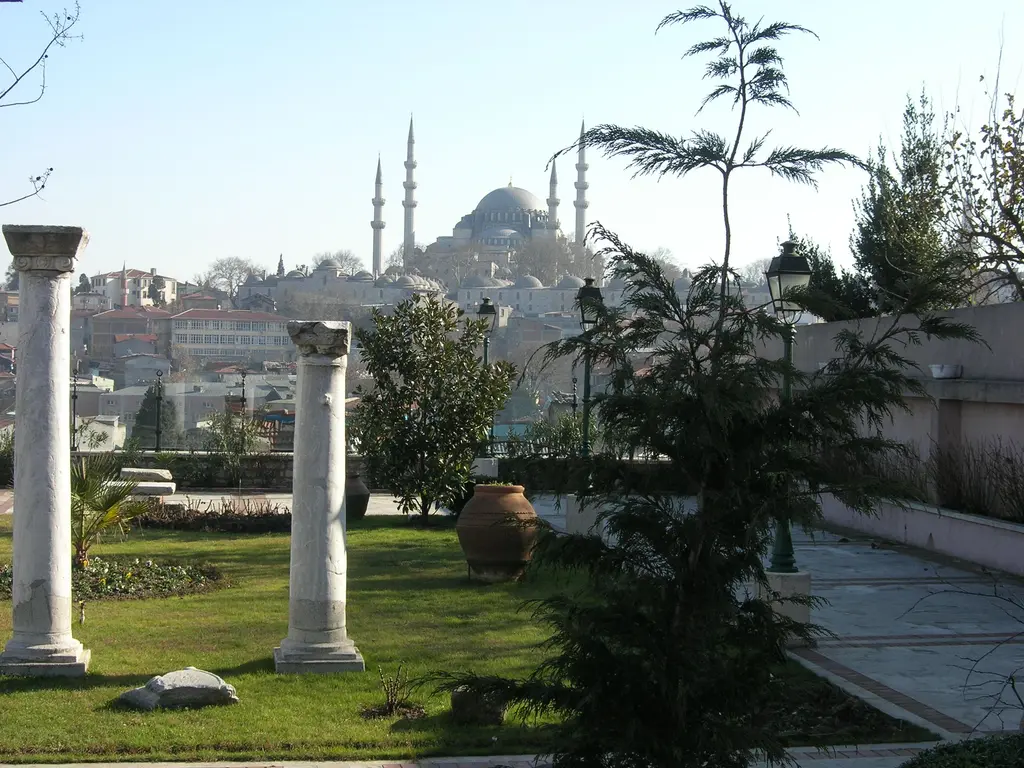 Zeyrek Camii'nin bahçesinden şehir manzarası; önde iki beyaz mermer sütun ile çim, ortada kiremit çatılı evler, uzakta tepede dört minareli Süleymaniye Camii.