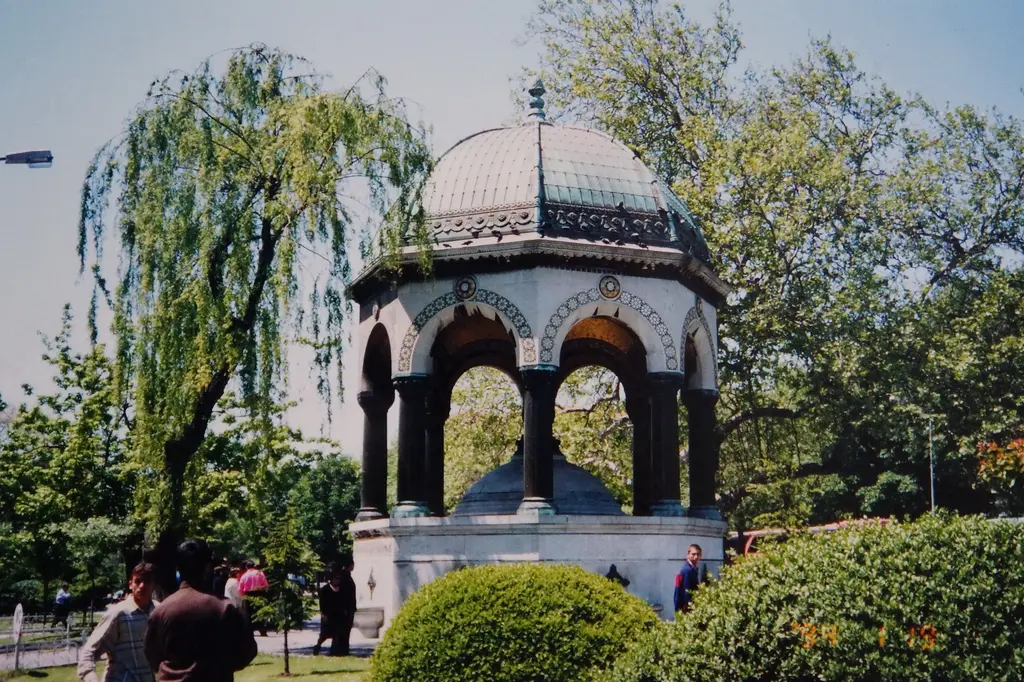 Ağaçlar arasında Alman Çeşmesi; gri kubbe, koyu sütunlar, önde yuvarlak budanmış çalılar, solda sarkan söğüt ve ağaç altında insanlar.