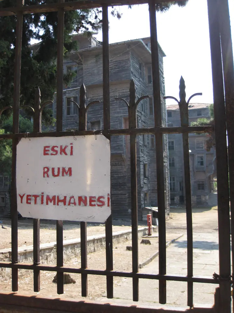 Büyükada Rum Yetimhanesi'nin kapısı; demir parmaklıklı kapı, üstünde "Eski Rum Yetimhanesi" yazılı tabela ve ardında ağaçlar.