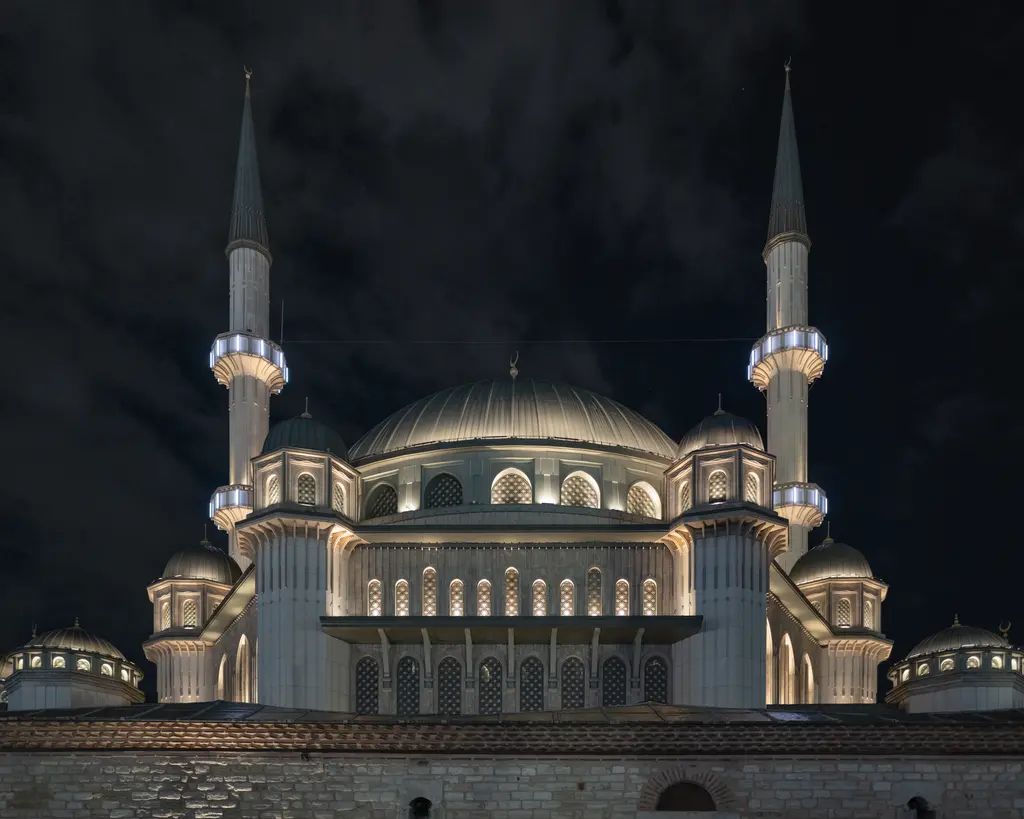 Taksim Camii gece; ışıklandırılmış krem cephe, sarı ışıkla aydınlanan kubbeler ve karanlık gökyüzüne yükselen iki minare.