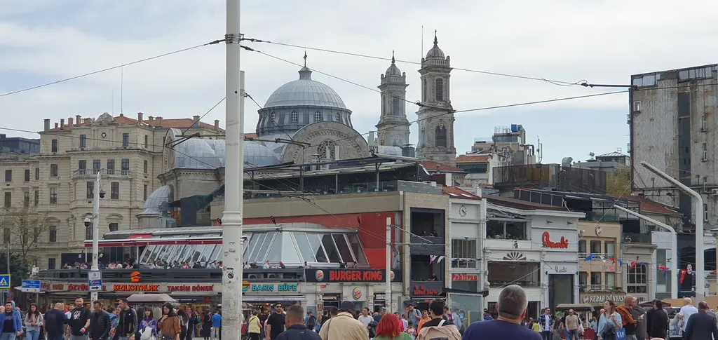 Taksim Meydanı'nda kalabalık; önde yürüyen insanlar ve dükkânlar, arkada Aya Triada Kilisesi'nin kubbesi ve iki çan kulesi.