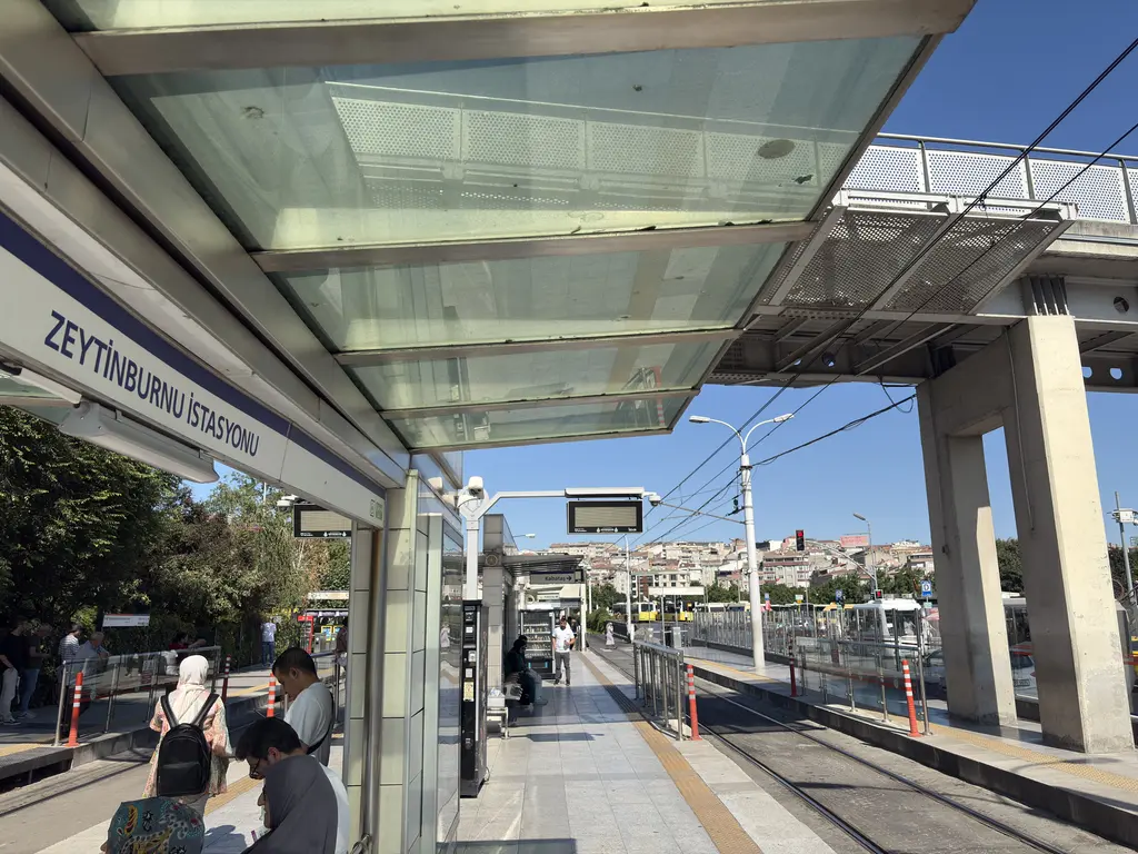Zeytinburnu tramvay istasyonunun peronu; cam saçak altında bekleyen yolcular, rayların üzerinde duran kırmızı beyaz tramvay ve üst geçit.