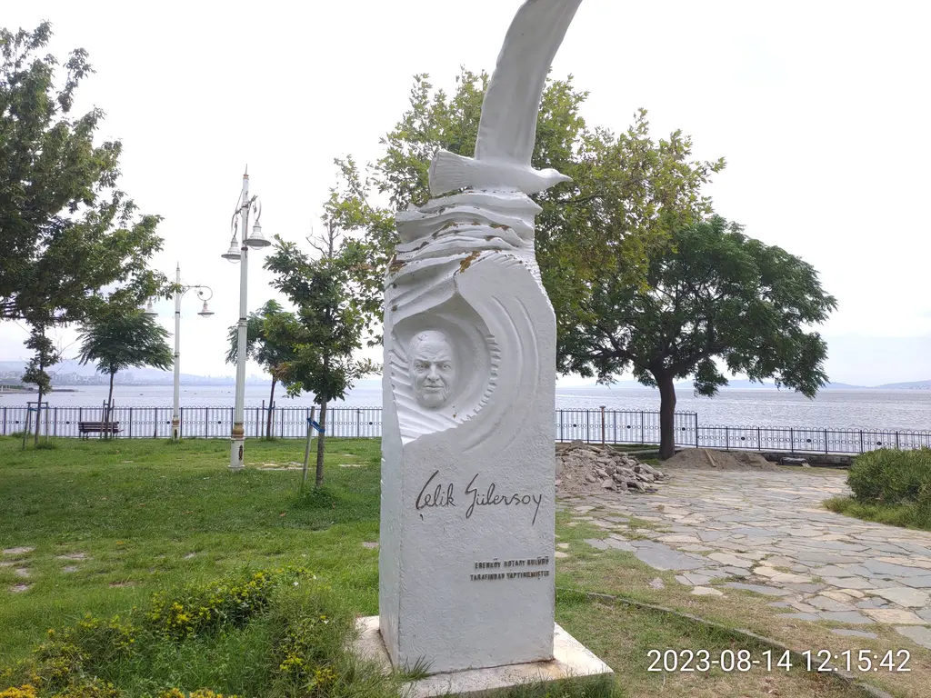 Fenerbahçe Parkı'nda Çelik Gülersoy Anıtı; beyaz mermerde kabartma portre ve imza, arkada ağaçlar ve deniz.
