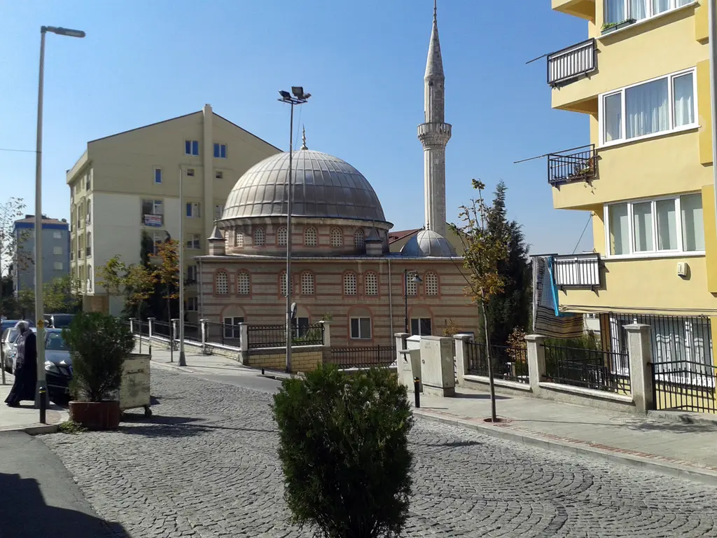 Kurtuluş'ta Hasan Zeynep Camii; tek kubbeli küçük cami ve ince minare, önde parke taşlı sokak, iki yanda apartmanlar.
