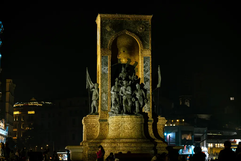 Gece sarı ışıkla aydınlatılmış Cumhuriyet Anıtı; kemerin içindeki bronz asker figürleri, iki yanda bayrak taşıyan heykeller.