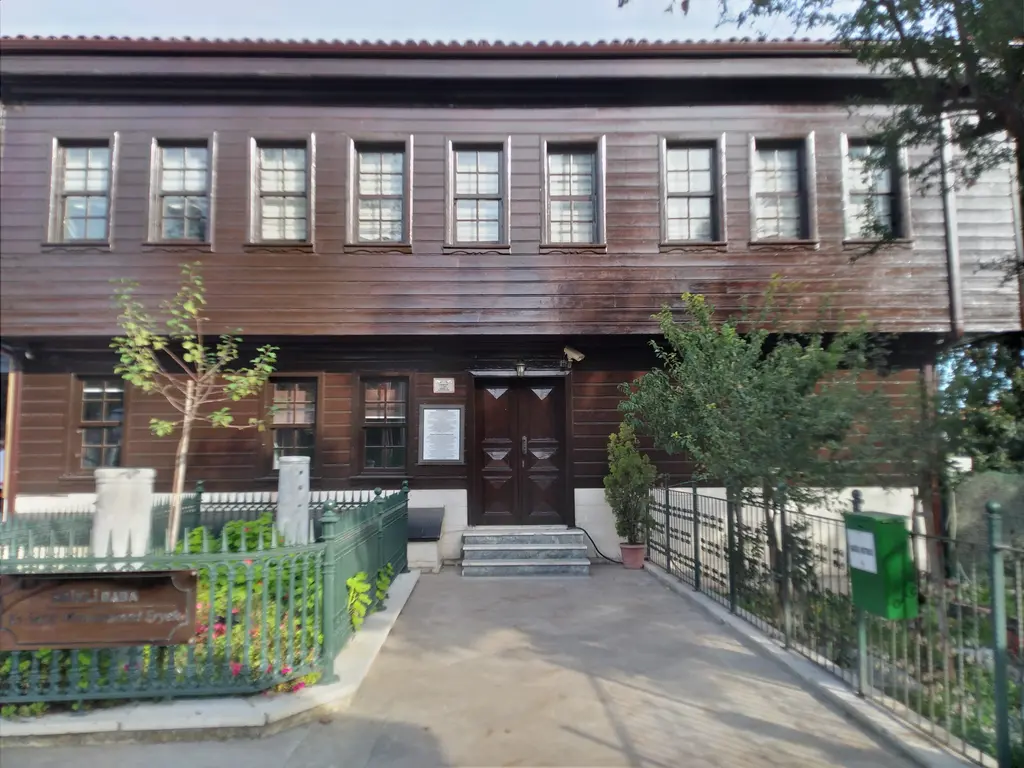 Erikli Baba Tekkesi; ahşap cepheli tek katlı yapı, kiremit çatı, önünde taş döşeli avlu ve saksılarda çiçekler.