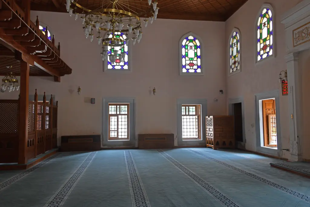 Şah Sultan Camii'nin harimi; ahşap mahfil, renkli camlı kemerli pencereler, avize ve mavi halı.