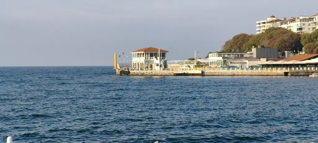 Moda İskelesi uzaktan; denizin ortasında beyaz bina, uzun iskele yolu ve arkada yamaçtaki apartmanlar.