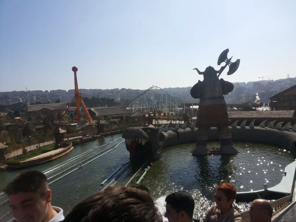 Vialand'de su havuzunun ortasında baltalı dev Viking heykeli; çevresinde su kanalı, arkada roller coaster rayları ve önde kalabalık.