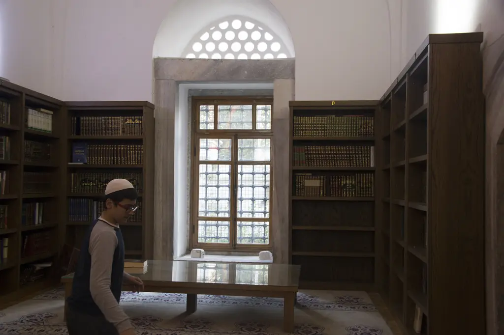 Yavuz Sultan Selim Camii'nde kuzey tabhane odası; duvar boyunca ahşap kitap rafları, önde masada çalışan bir okur ve dilimli kemerli pencere.