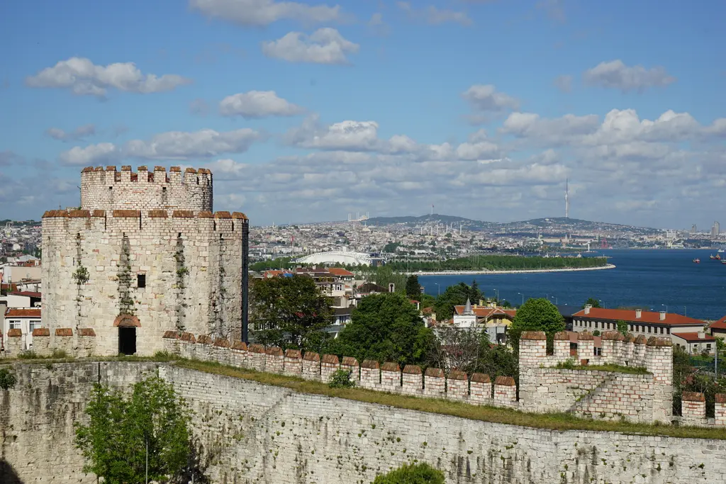 Yedikule Hisarı'nda duvarın üzerinden yükselen kırmızı kiremit başlıklı yuvarlak mazgallı kule; arkada şehrin çatıları, yeşil park, Marmara Denizi ve pamuk bulutlu mavi gökyüzü.