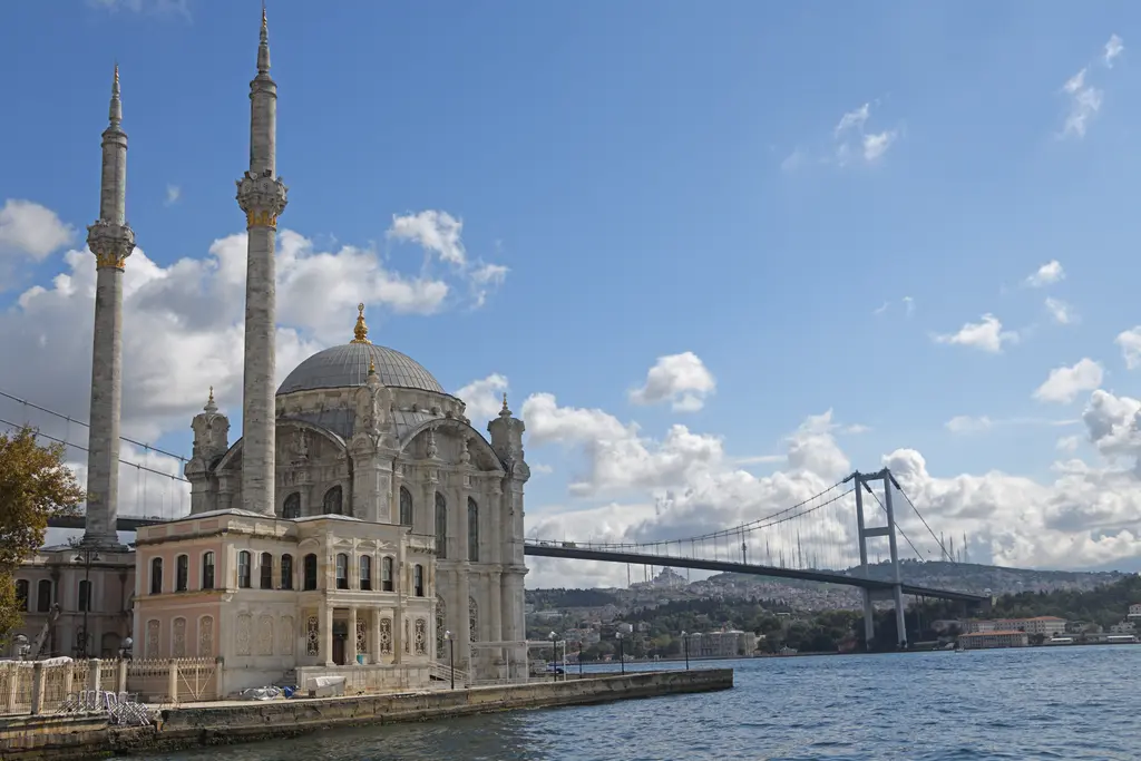 Beşiktaş'ta Ortaköy Camii; kıyıdaki beyaz taş cami, iki ince minare, arkada Boğaz'ı aşan asma köprü ve bulutlu mavi gökyüzü.