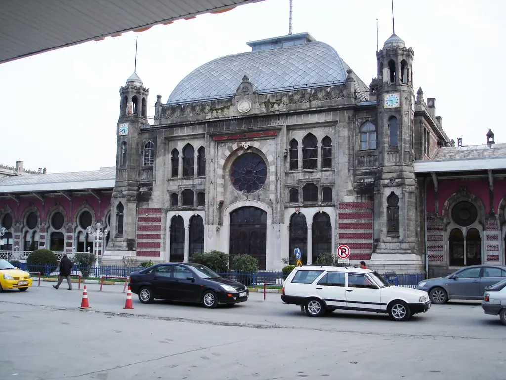 Sirkeci Garı'nın ana cephesi karşıdan; ortada büyük gül pencere ile kemerli giriş, iki yanda ince kuleler, üstte dilimli cam kubbe ve caddede park etmiş araçlar.