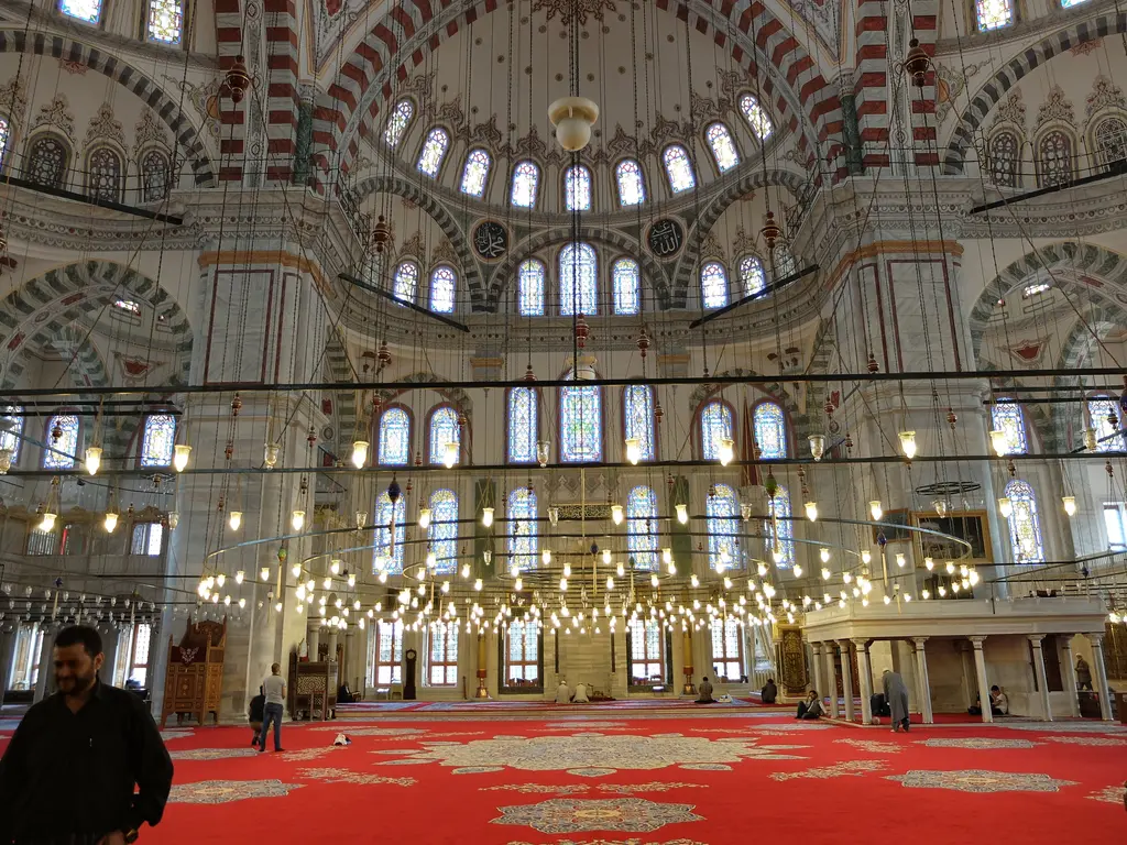 Fatih Camii'nin harimi mihrap yönüne bakışla; beyaz gri duvarlar, kırmızı beyaz almaşık kemerler, kat kat vitraylı pencereler, alçak kandil halkası ve madalyonlu kırmızı halı.