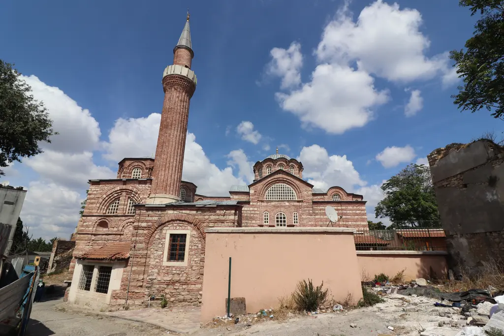 Vefa Kilise Camii'ne doğudan bakış; bahçe duvarının üstünde yivli tuğla minare, kubbe kasnağı, büyük kemerli alınlık penceresi ve sağır kemer dizisi, önde moloz ve otlar.