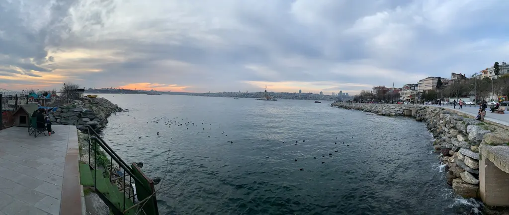 Salacak sahili; bulutlu gökyüzü altında taş dolgu kıyı, deniz kenarında yürüyenler ve uzakta tarihi yarımada.