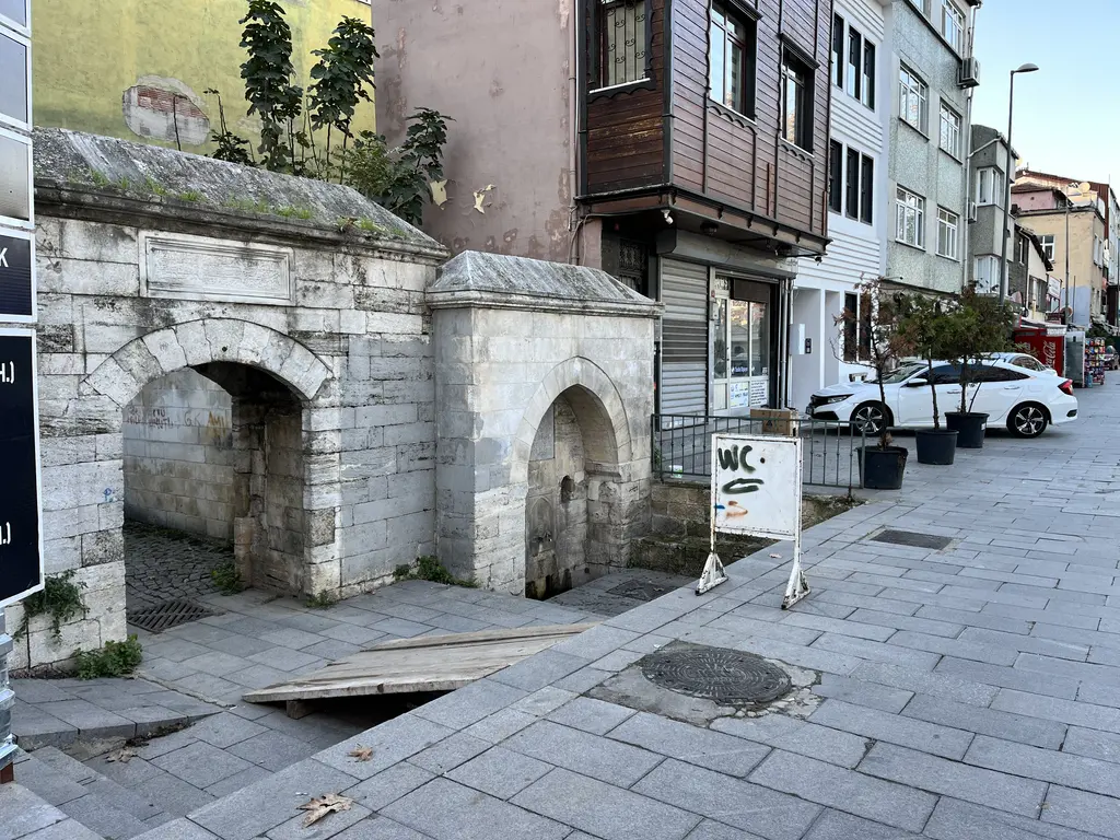 Eyüp'te Defterdar Çeşmesi ve sokak; taş çeşme, kemerli kapı, yanında ahşap cumbalı ev, park etmiş araçlar ve kaldırım.