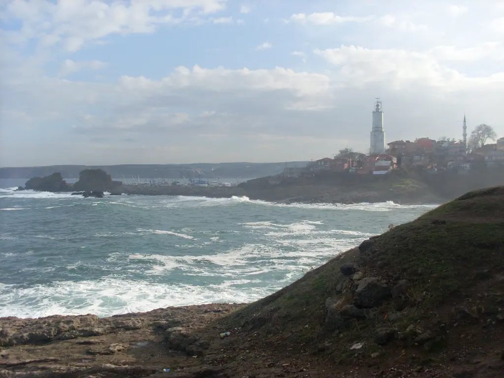 Rumelifeneri'nde Karadeniz; köpüklü dalgalar, kayalık burun, karşıda beyaz deniz feneri, köy evleri ve minare.