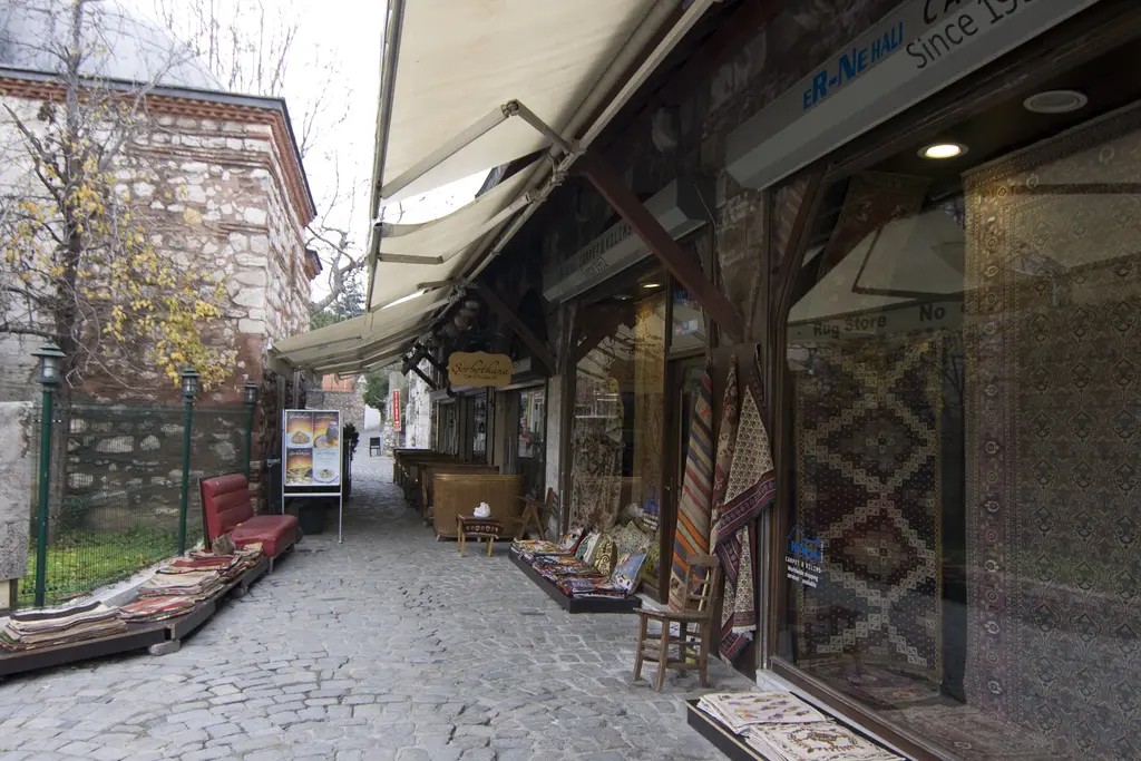 Arasta Çarşısı'nın ucu; solda ağaçlar ve demir parmaklık, sağda kemerli dükkânlar, halı vitrinleri ve taş döşeli yol.