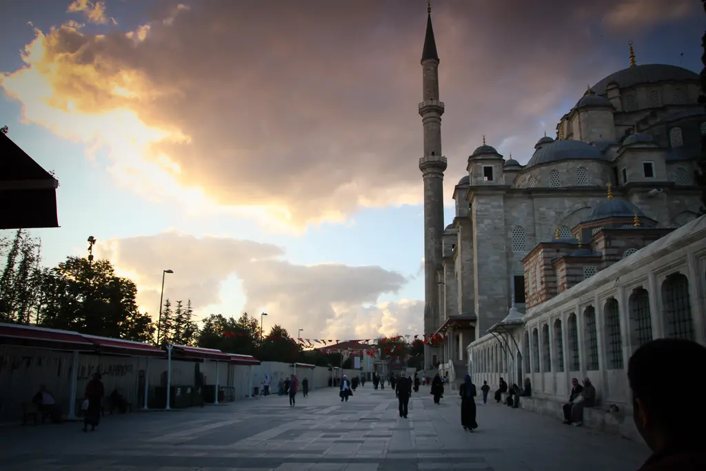 Fatih'te gün batımında Fatih Camii; gökyüzünü dolduran turuncu altın bulutlar, sağda caminin silueti ile minaresi ve taş döşeli meydanda yürüyen insanlar.