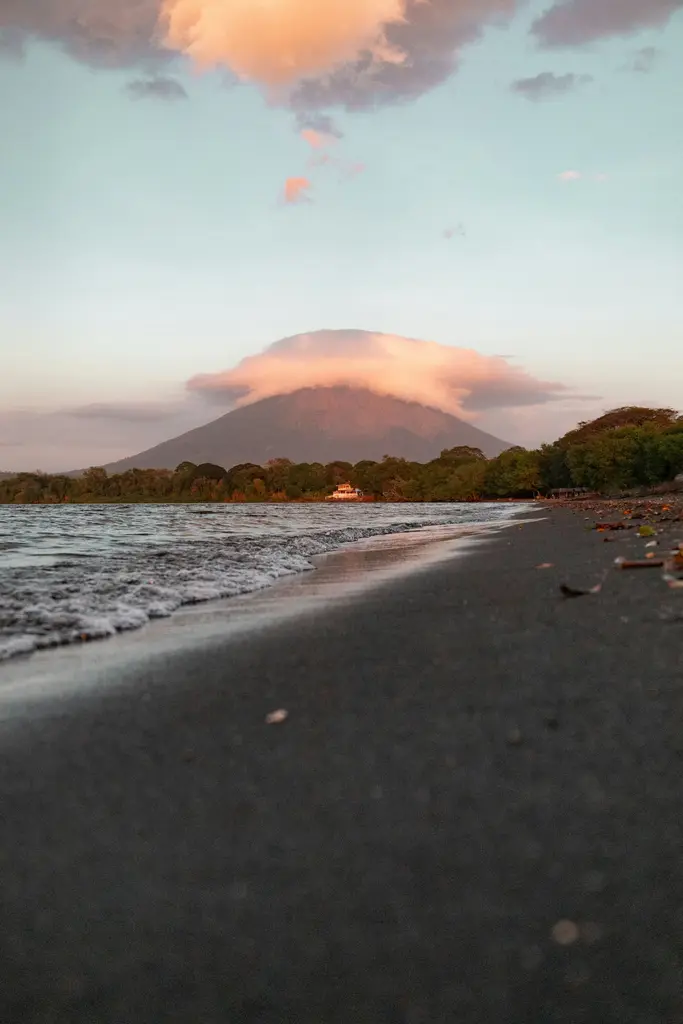 Nikaragua'da Ometepe Adası'nda gün batımında zirvesi bulutla örtülü Concepción Yanardağı; önde göl kıyısının koyu kumlu plajı ve kıyıya vuran dalgalar.