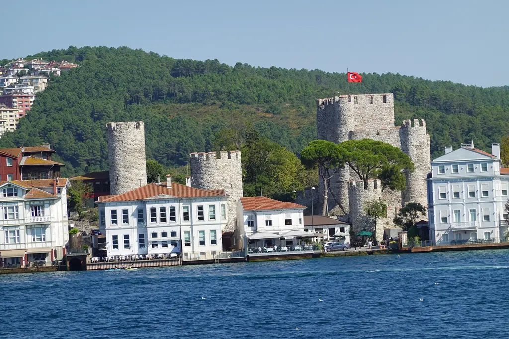 Beykoz'da Anadolu Hisarı; Boğaz kıyısında bayraklı iç kale burcu, yuvarlak kuleler, önünde beyaz yalılar ve deniz.