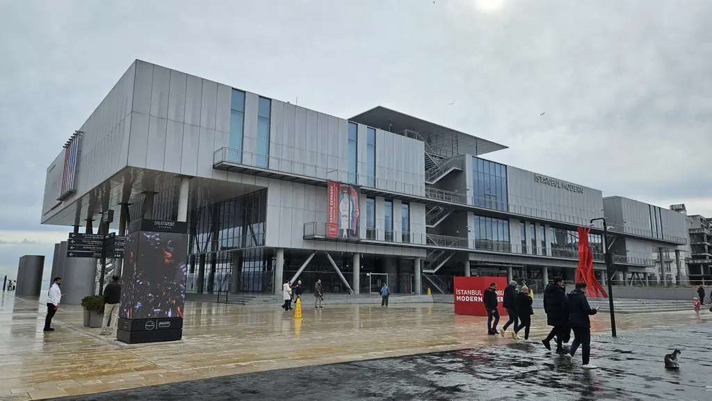 Yağmurlu bir günde İstanbul Modern; ıslak meydanda yansımalar, kırmızı tabelalar ve binanın çapraz açıdan görünen kütleleri.