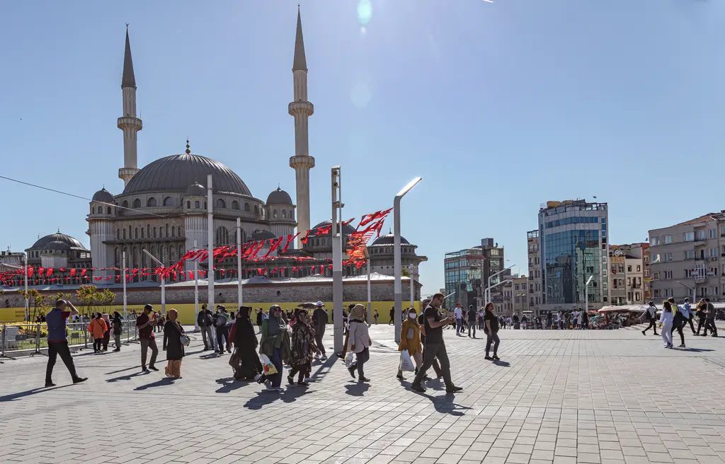Açık bir günde Taksim Camii; önündeki geniş meydanda yürüyen kalabalık, sağda çok katlı binalar ve gerili flamalar.