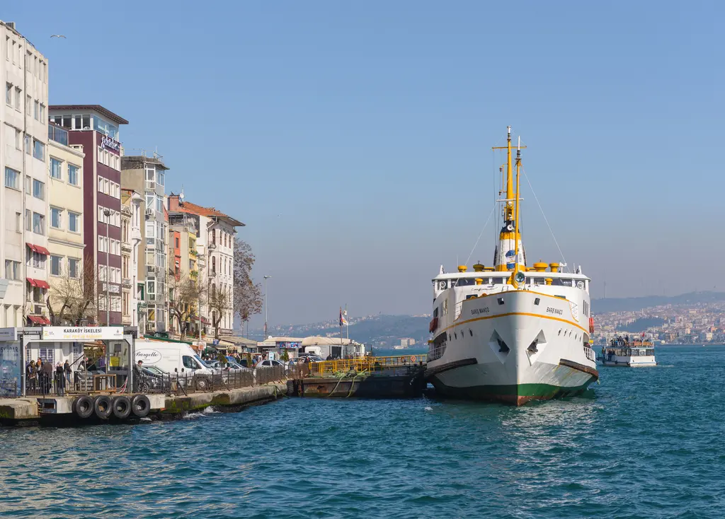 Karaköy'de iskeleye bağlı vapur; sarı bacalı beyaz gemi, rıhtımdaki park etmiş araçlar ve solda yüksek binalar.