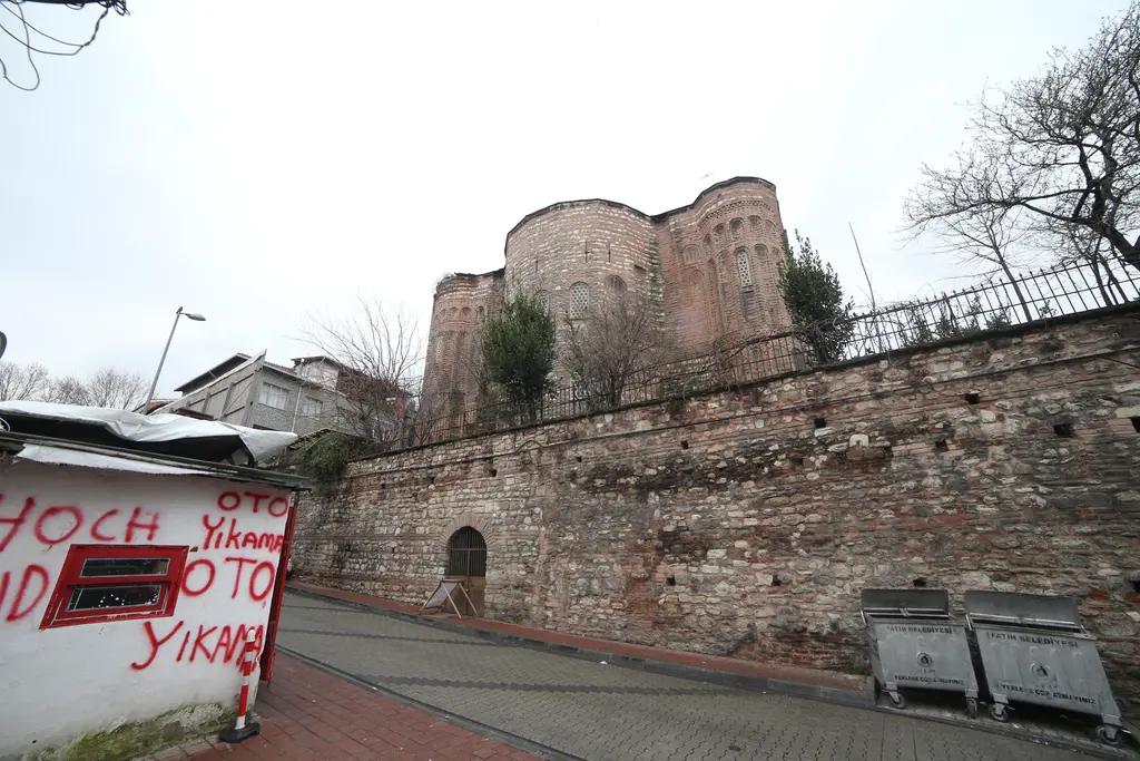 Gül Camii'nin kubbeleri ve önünde uzun taş duvar; sokakta çöp konteynerleri ve sol köşede yazılı duvar.